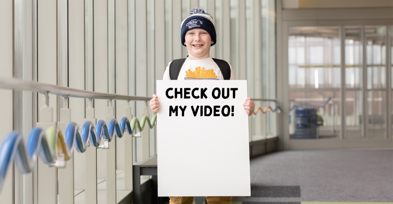 David, 2026 KIDstruction Week patient ambassador, holding a sign that reads CHECK OUT MY VIDEO
